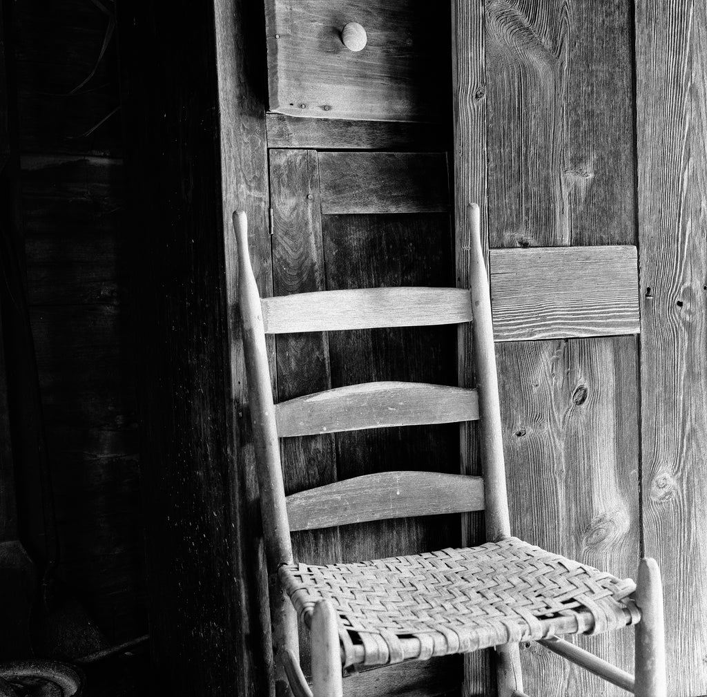 Black and white photograph of an old wooden chair inside a rustic mountain cabin in the American South. This photograph was shot on medium format Kodak TMAX 100 black and white film. Film photography provides a different look than digital photography and prints will display a pleasing amount of film grain and may appear somewhat softer than modern digital images. (Square format)