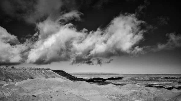 Black and white panoramic landscape photograph of a dramatic low-hanging cloud casting a shadow over the hills of the Painted Desert in Arizona.