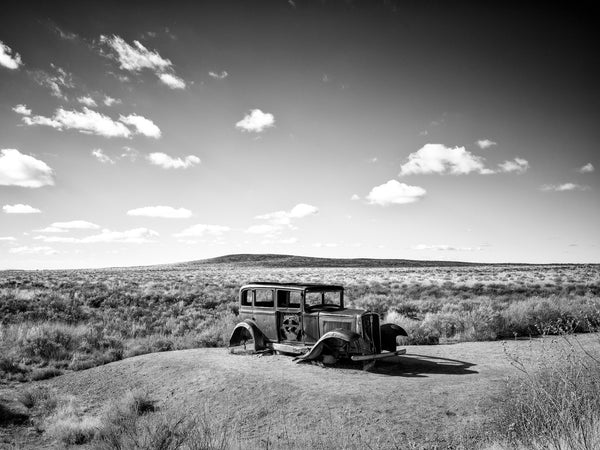 Black and white photograph of a rusty antique car on an old alignment of Route 66 in the boundaries of the Petrified Forest in Arizona.