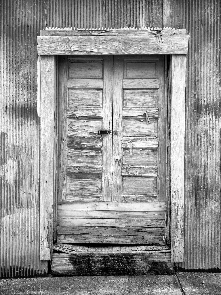 Black and white photograph of textured and weathered wooden doors on the corrugated steel wall of a big cotton warehouse in the American South.