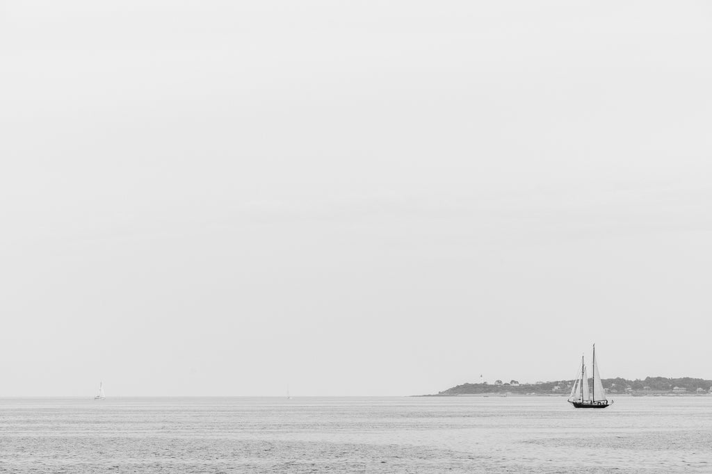 Black and white photograph of sail boats cutting through sun-washed haze of the New England coast with a lighthouse visible in the distance.