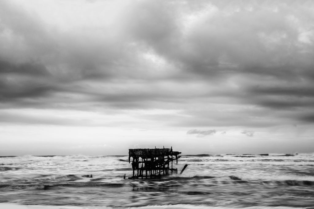 Black and white photograph of the rusty bones of the wreck of the Peter Iredale on the stormy coast of Oregon near Astoria. The metal ship ran aground during strong winds in 1906 with no injuries. Normally it's visible in the sand, but on this morning the rough tide washed over it.