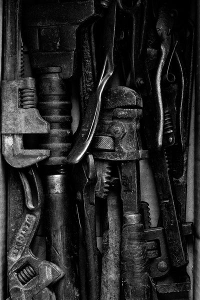 Black and white photograph of a box containing a pile of antique hand tools shot in dramatic light to accentuate the textures and details of the rugged old steel and wood.