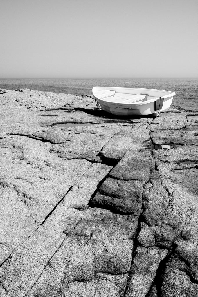 Black and white photograph of a small boat perched on the rocky sea coast of Maine overlooking the vast Atlantic Ocean.