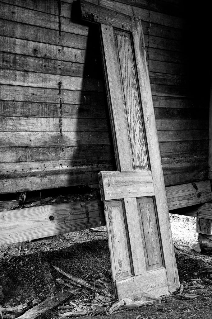 Black and white photograph of the broken old front door of an abandoned 1890s farmhouse now leaning against a wall inside the structure.