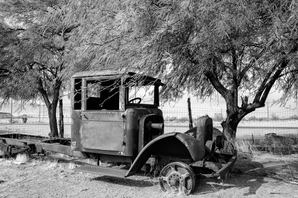 Black and white photograph of the skeletal remains of a rusty old truck parked alongside the dusty stretch of old Route 66 that runs through the Sitgreaves Pass to Oatman, Arizona.