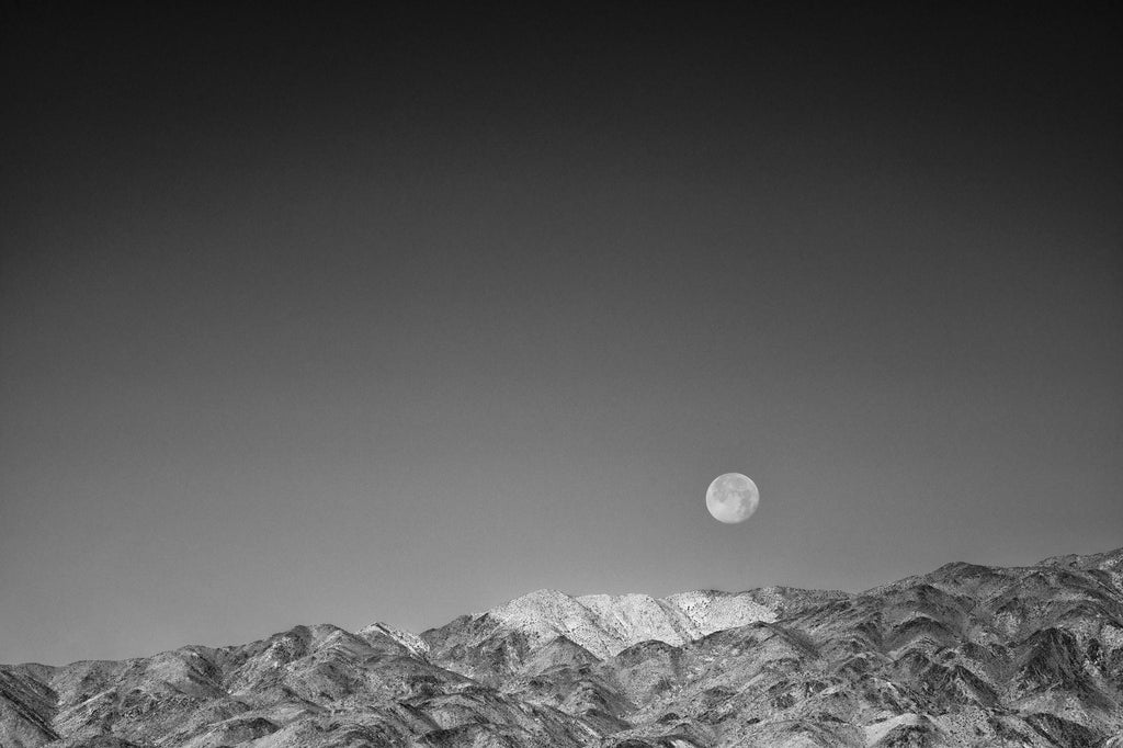 Black and white photograph of a low moon over the mountains at Joshua Tree National Park in California.