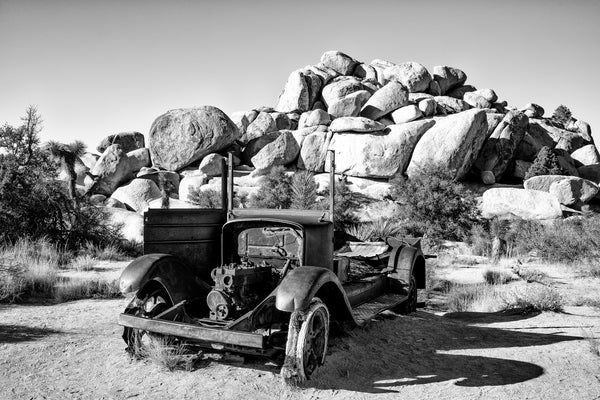 Black and white landscape photograph of a rusty old abandoned truck sitting in the desert near a rock formation at Joshua Tree National Park in California.