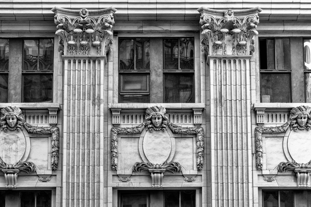Black and white photograph of a building in downtown Los Angeles with ornate architectural details including a row of female heads wearing feather headdresses and necklaces with large round beads.