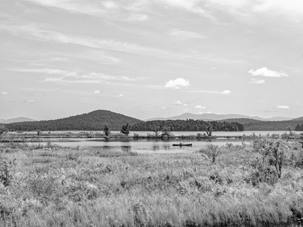 Black and white photograph of a kayaker traversing a pond in the beautiful landscape of upstate New York's Adirondack Mountains.
