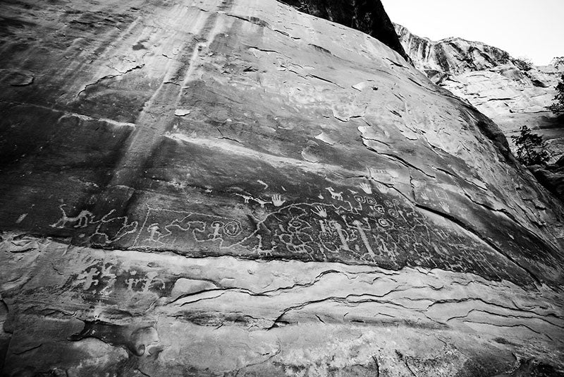 Black and white fine art photograph of ancient petroglyphs that can still be seen at Mesa Verde, Colorado. Petroglyphs are images made by scratching or carving into stone. These glyphs tell the origin story of the Pueblo people. This photograph makes the glyphs appear high on the wall, but in actuality, the glyphs were within reach using a narrow stone ledge just below the wall.
