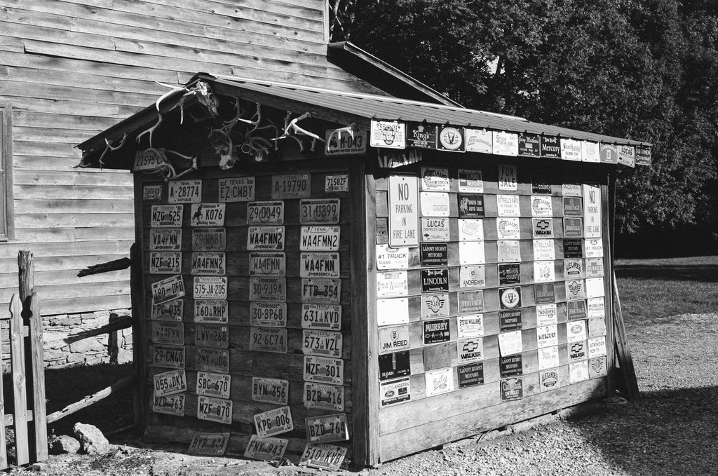 Black and white photograph of an old wooden shed covered with old license plates and crowned with antlers. This photograph was shot on 35mm Kodak Tri-X black and white film using a vintage Pentax camera. Film photography provides a different look than digital photography and prints will display a pleasing amount of film grain and may appear somewhat softer than modern digital images.