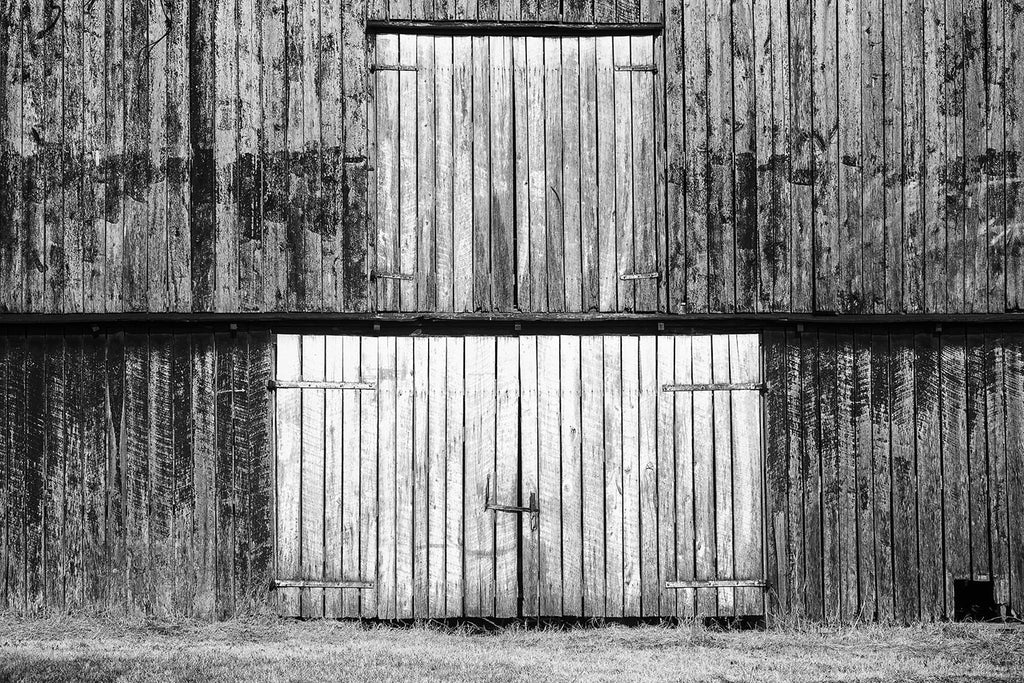 Black and white photograph of barn doors on the first and second levels of a big old weather-beaten red barn. This photograph is composed flatly, allowing the rectangles and vertical lines to create an almost abstract architectural image.