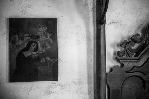 Black and white photograph of an antique portrait of The Madonna in Nun's habit hanging on a wall near the post of a wooden bed inside an old house in Lafayette, Louisiana. 