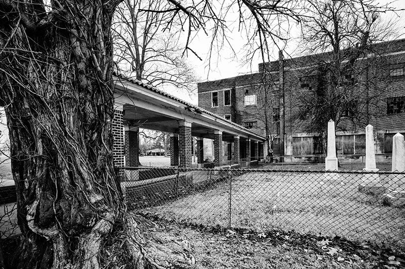 Black and white photograph of a very large abandoned school building in Clarksdale, Mississippi, with the remains on an old family cemetery on the school yard. NOTE: I had originally, mistakenly called this a confederate cemetery, but it's actually the Bobo family cemetery, with headstones ranging from 1801 to 1895.