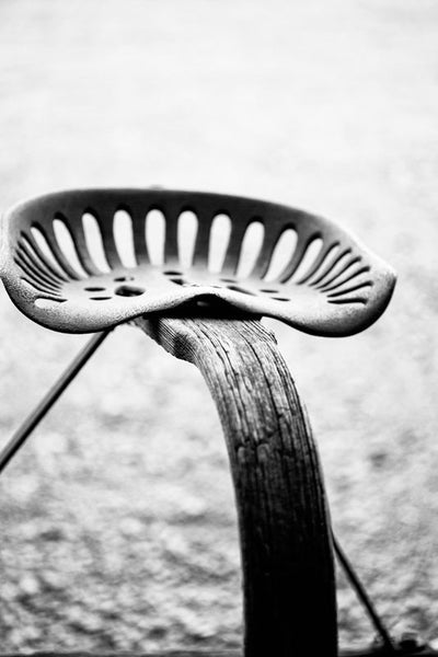 Black and white photograph of a rusty metal plow seat on a weathered wooden stand, part of an antique farm machine.