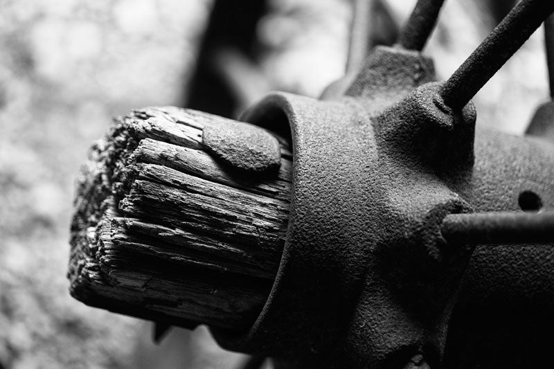 Black and white photograph of a rusty antique spoked metal wheel with a rough wooden axle. This may have been part of a piece of farm equipment.