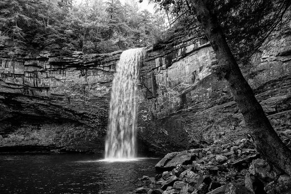 Black and white landscape photograph of a waterfall in soft light, emphasizing the whitewater of the river spilling over the edge of the cliff, and the high rocky walls of the basin that surrounds the pool at the base of the falls.