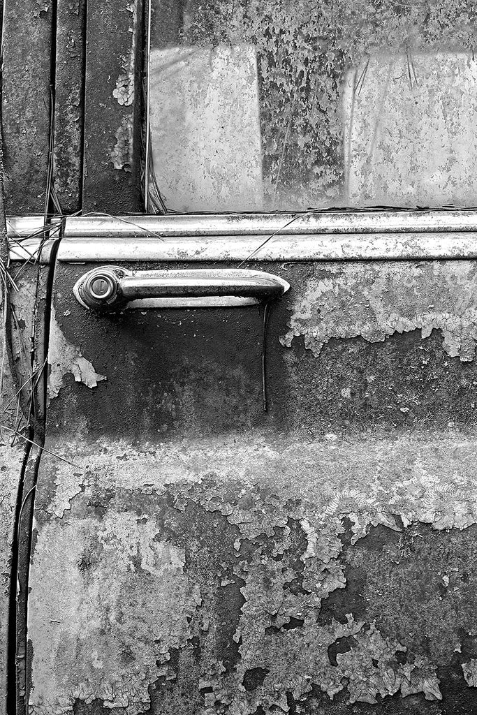 Black and white fine art detail photograph of rust and peeling paint on the door of an junked classic American car.