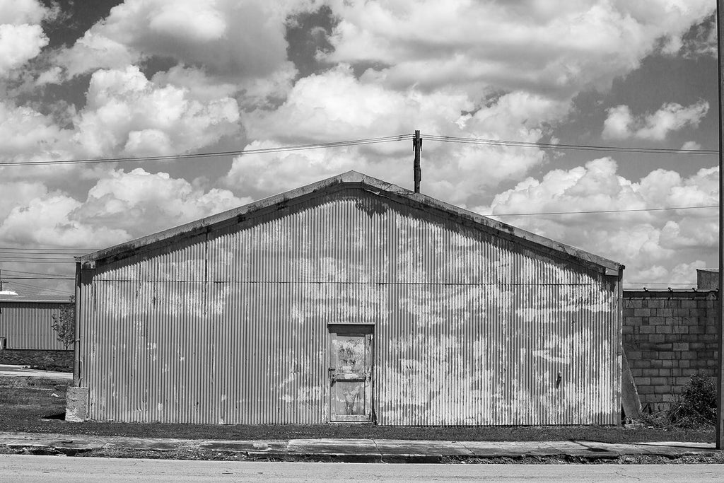 Black and white photograph of a tin industrial building with vertical ridges pointing up to a beautifully cloudy sky.