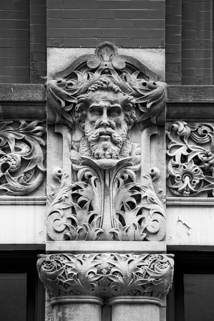 Black and white photograph of a carved face on the Drhumor Building (pronounced "drummer") on 48 Patton Avenue in downtown Asheville, North Carolina. Built in 1895, the building features a set of carved faces, believed to have been modeled by local citizens, created by English sculptor Frederick Miles, who also worked on the nearby Biltmore Estate. This photograph is one of a set of six, designed to be displayed together.