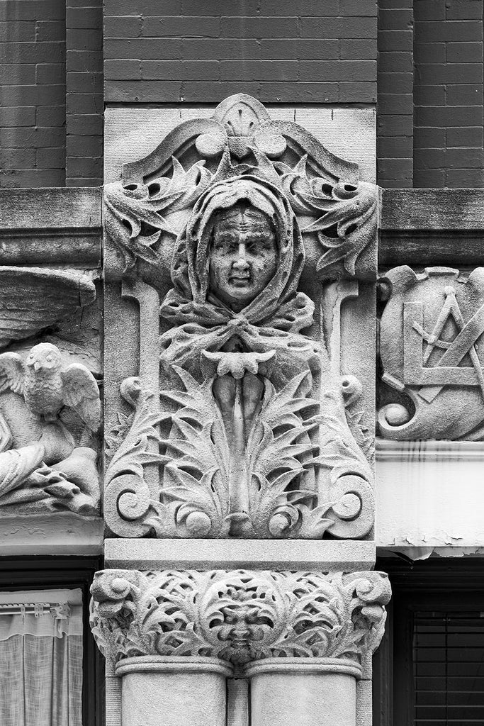 Black and white photograph of a woman's face carved on the Drhumor Building (pronounced "drummer") on 48 Patton Avenue in downtown Asheville, North Carolina. Built in 1895, the building features a set of carved faces, believed to have been modeled by local citizens, created by English sculptor Frederick Miles, who also worked on the nearby Biltmore Estate. This photograph is one of a set of six, designed to be displayed together.