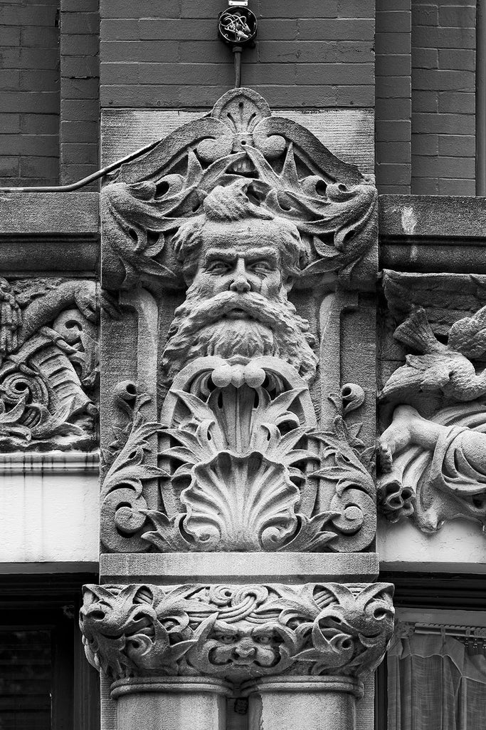 Black and white photograph of a bearded man's face carved on the Drhumor Building (pronounced "drummer") on 48 Patton Avenue in downtown Asheville, North Carolina. Built in 1895, the building features a set of carved faces, believed to have been modeled by local citizens, created by English sculptor Frederick Miles, who also worked on the nearby Biltmore Estate. This photograph is one of a set of six, designed to be displayed together.