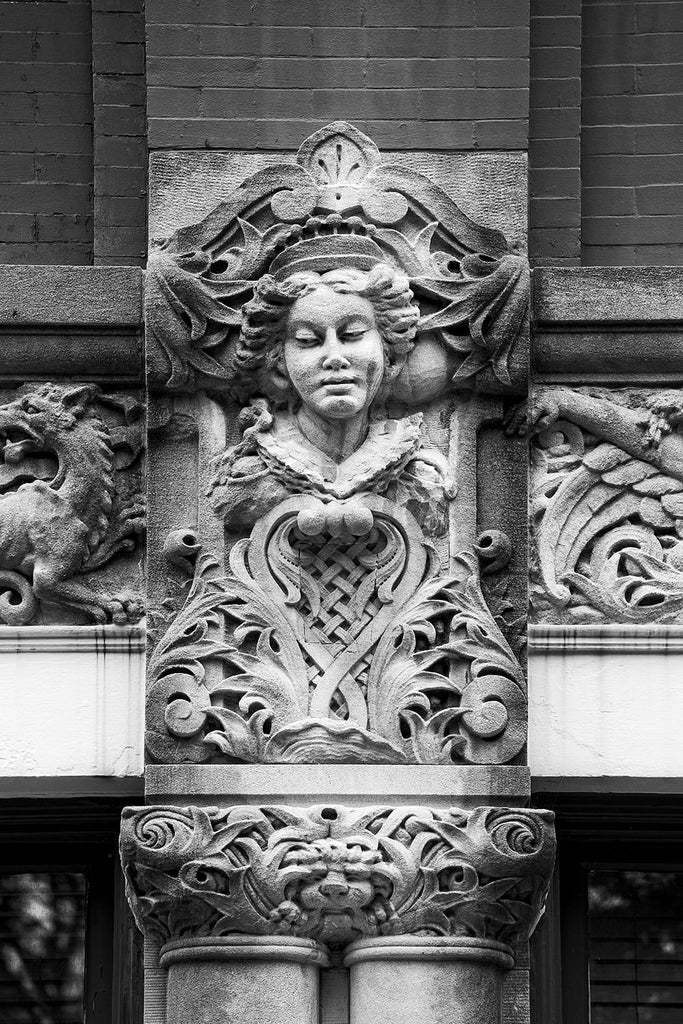 Black and white photograph of a young woman's face carved on the Drhumor Building (pronounced "drummer") on 48 Patton Avenue in downtown Asheville, North Carolina. Built in 1895, the building features a set of carved faces, believed to have been modeled by local citizens, created by English sculptor Frederick Miles, who also worked on the nearby Biltmore Estate. This photograph is one of a set of six, designed to be displayed together.