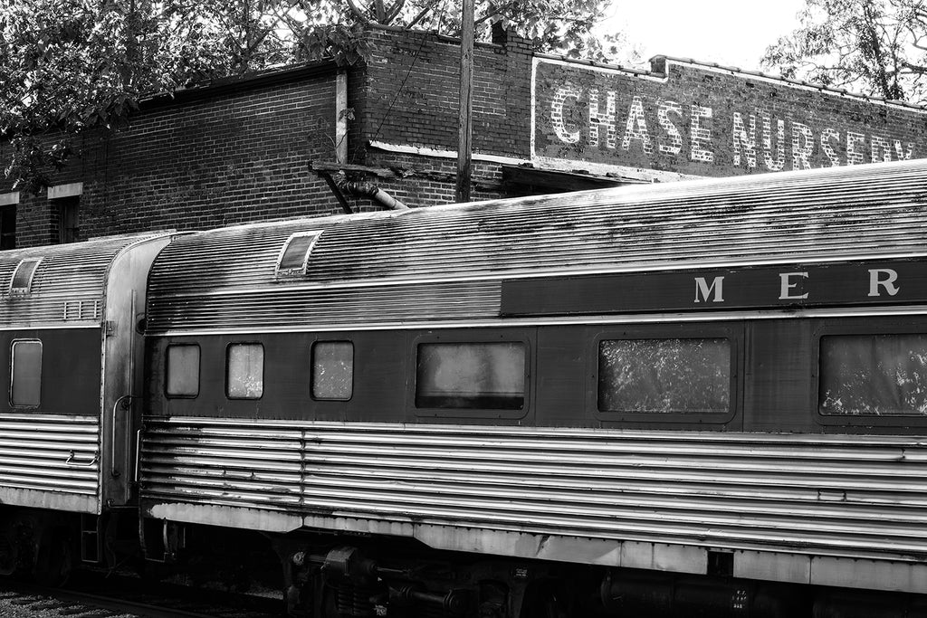 Black and white photograph of a rusty set of retired railroad passenger cars parked on decommissioned railroad tracks outside an abandoned station.