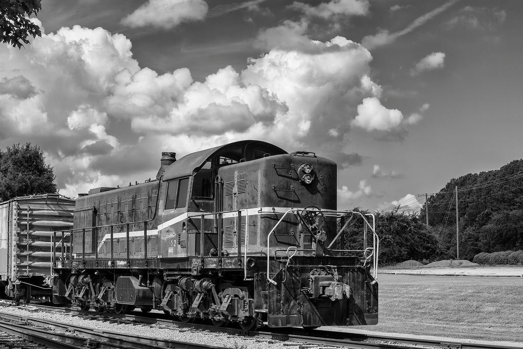 Black and white photograph of an retired old train locomotive sitting on railroad tracks among the trees.