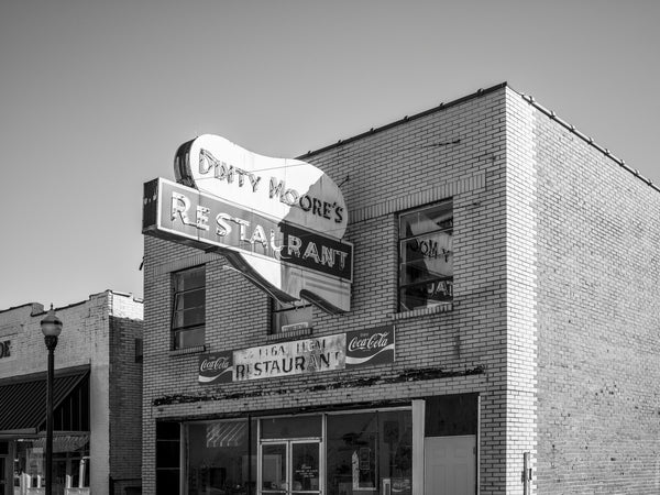 Black and white photograph of the abandoned storefront and vintage sign for a Dinty Moore Restaurant in a small southern town. Subsequent businesses left the sign intact but the building is now vacant.