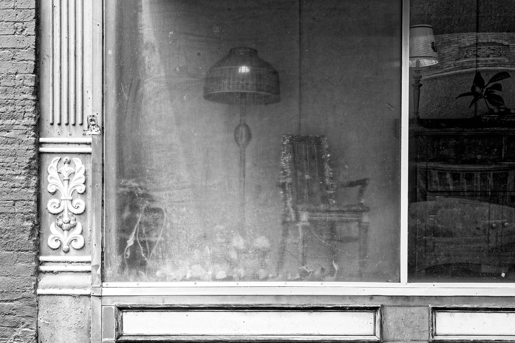 Black and white photograph of old furniture arranged behind hazy glass in the window display of a main street shop in a small town.
