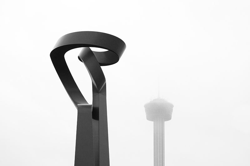 Black and white photograph of two of San Antonio's most iconic landmarks on a foggy morning -- San Antonio's big, red sculpture "La Antorcha de la Amistad" (The Torch of Friendship) by Mexican artist Sebastian, and in the distance, shrouded in fog, is Tower of the Americas.