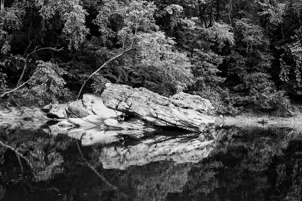 Black and white landscape photograph of large boulders reflecting into the placid surface of a slow-moving river.