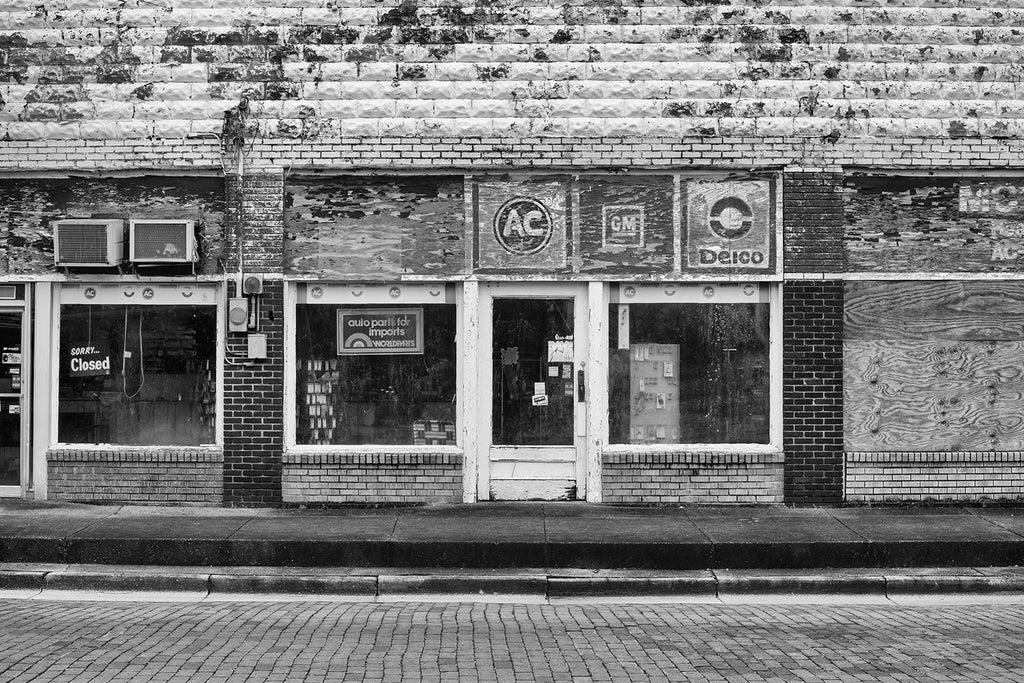 Black and white architectural detail photograph of an apparently defunct auto parts storefront in a small town. Every quadrant of this photograph is rich with textures.