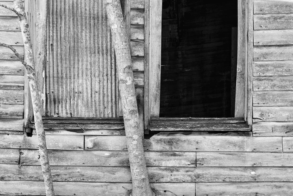 Black and white photograph of two windows on the side of an abandoned old clapboard house. One of the windows is covered in corrugated tin, while the other is wide open and dark.