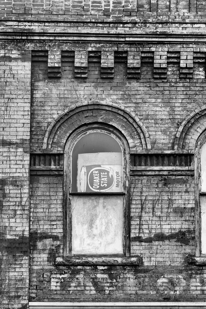 Black and white photograph of an arched second floor storefront window with a sideways motor oil sign and faded paint that says "Taylor" on dark bricks.