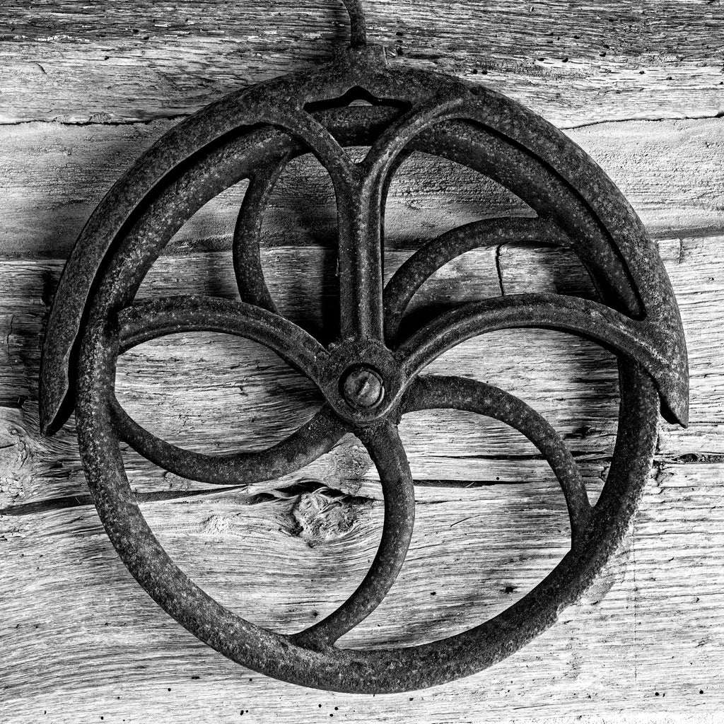 Black and white photograph of a rusty antique pulley wheel on an old farm, may have been used to pull buckets from a well.