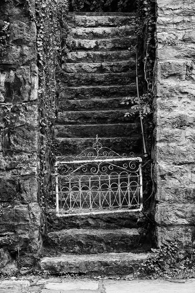Black and white photograph of a small, white, antique gate at the bottom of steep stone steps in a small town.