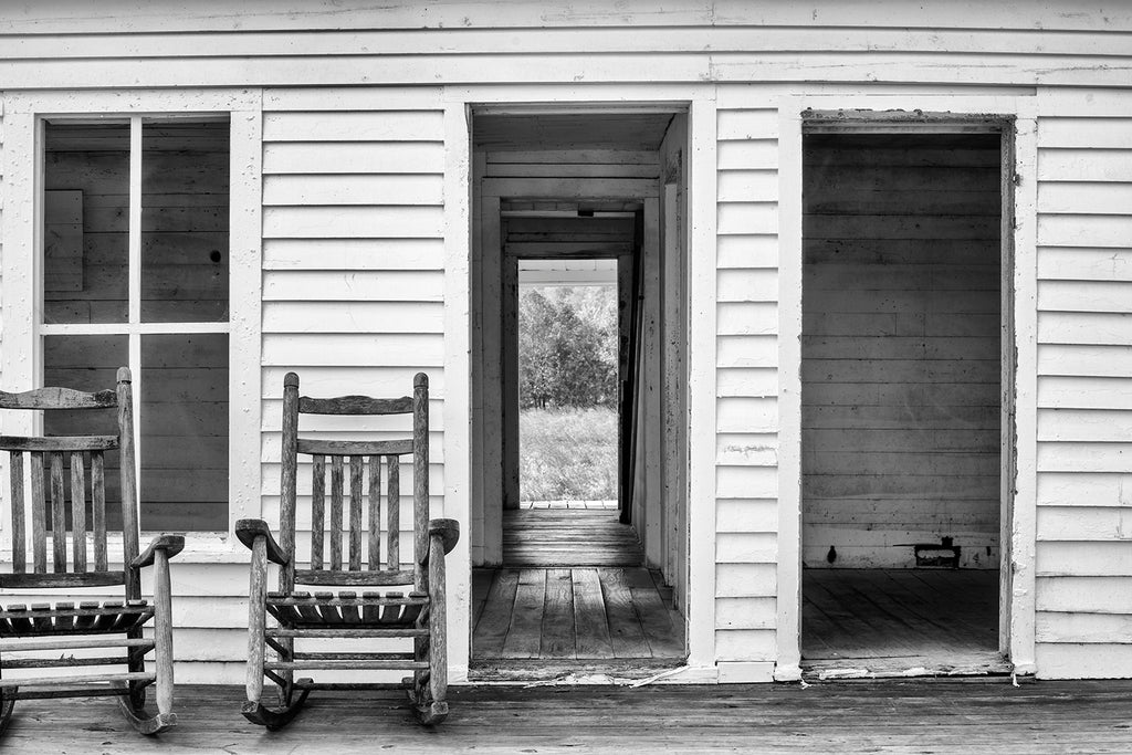 Black and white photograph of an abandoned farm house with rocking chairs on the porch.