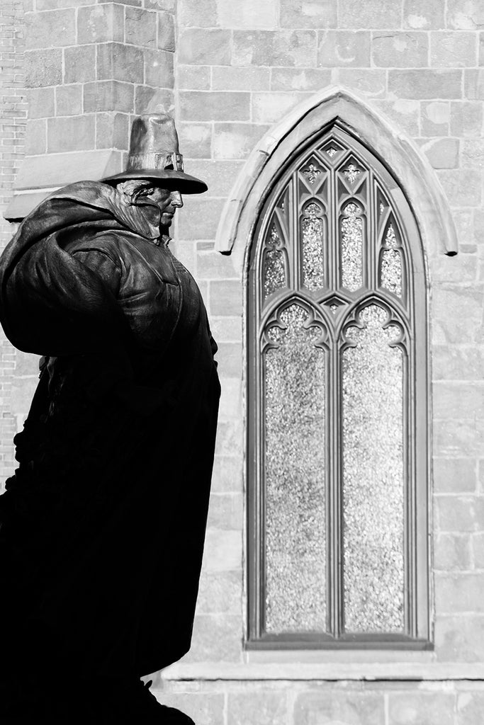 Black and white photograph of the statue of town founder Roger Conant by artist Henry Hudson Kitson, seen in the early morning shadows as the sun rises on Salem, Massachusetts. Behind the statue is a window of a former church that now holds a museum.