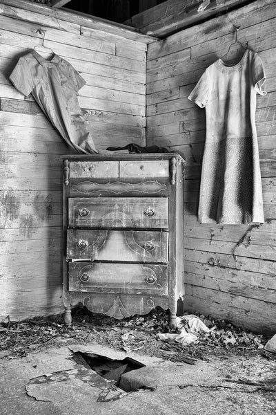 Black and white photograph of the interior of an abandoned and collapsing old house in the country with furniture left behind and dresses hanging on the walls.