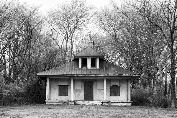 Black and white photograph of the front facade of an abandoned house with four posts on the front porch.