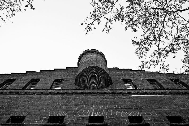 Black and white photograph of University of Wisconsin's Armory and Gymnasium, in Madison, Wisconsin. Better known as the old red gym, the structure was designed in the Romanesque revival style resembling a red brick castle. It was originally used as a combination gymnasium and armory beginning in 1894. It may seem odd to combine a gym with an armory, but it was funded after the Chicago Haymarket Riot in a time of labor unrest, when cities were building militias to put down worker strikes and protests.
