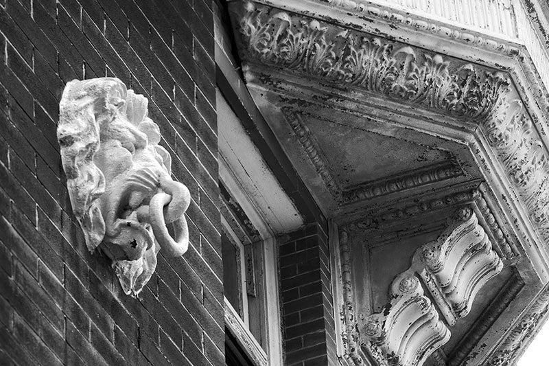 Black and white fine art photograph of architectural details along New Orleans' famous Canal Street, including a lion head with a tie-down ring in its mouth, and an ornamental pressed metal facade with peeling paint.