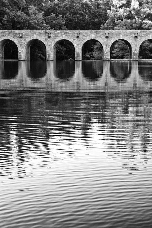 Black and white landscape photograph of the arches of an old stone bridge reflecting in quiet water.