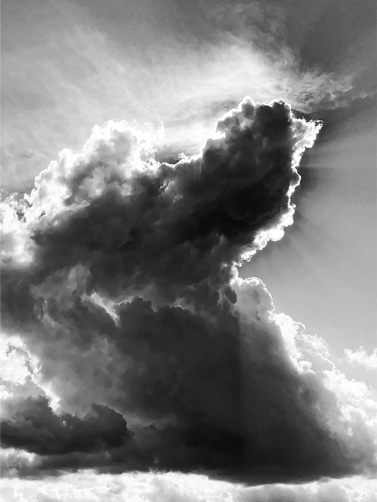 Black and white photograph of the sun glowing behind the boiling expanse of a tall developing storm cloud.