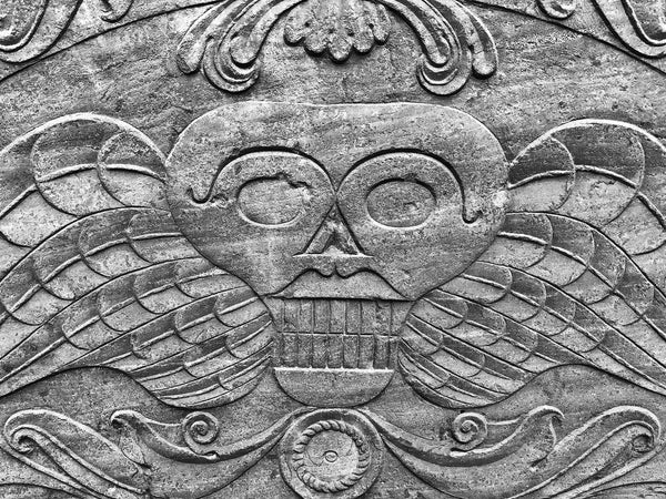 Black and white photograph of an iconic winged skull tombstone design commonly found on slate gray headstones of the 1700s. This particular design is located on a mid-1700s stone in Charleston.