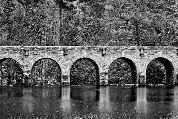 Black and white landscape photograph of an old stone bridge reflecting in a cold river in winter, with snowflakes flying.