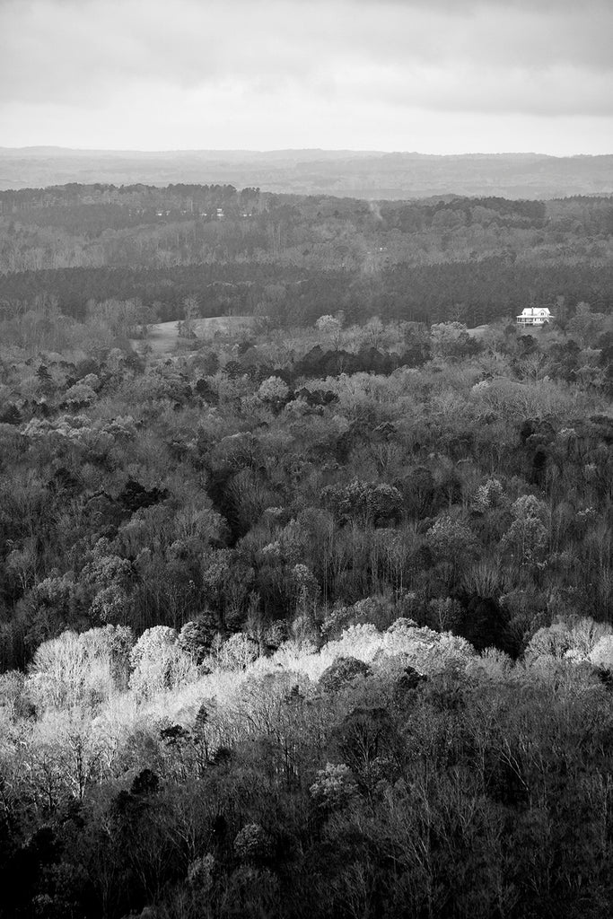 Black and white landscape photograph showing a bright shaft of sunlight crawling slowly across the forested floor of a valley with a white farmhouse in the distance.